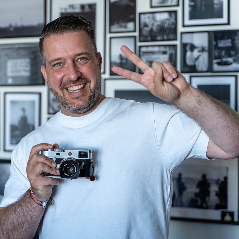 Maik Taubitz Bild zeigt Maik Taubitz in einer freundestrahlenden Pose. In einer Hand seine Leica-Kamera, mit der anderen Hand zeigt er das Victory-Zeiche